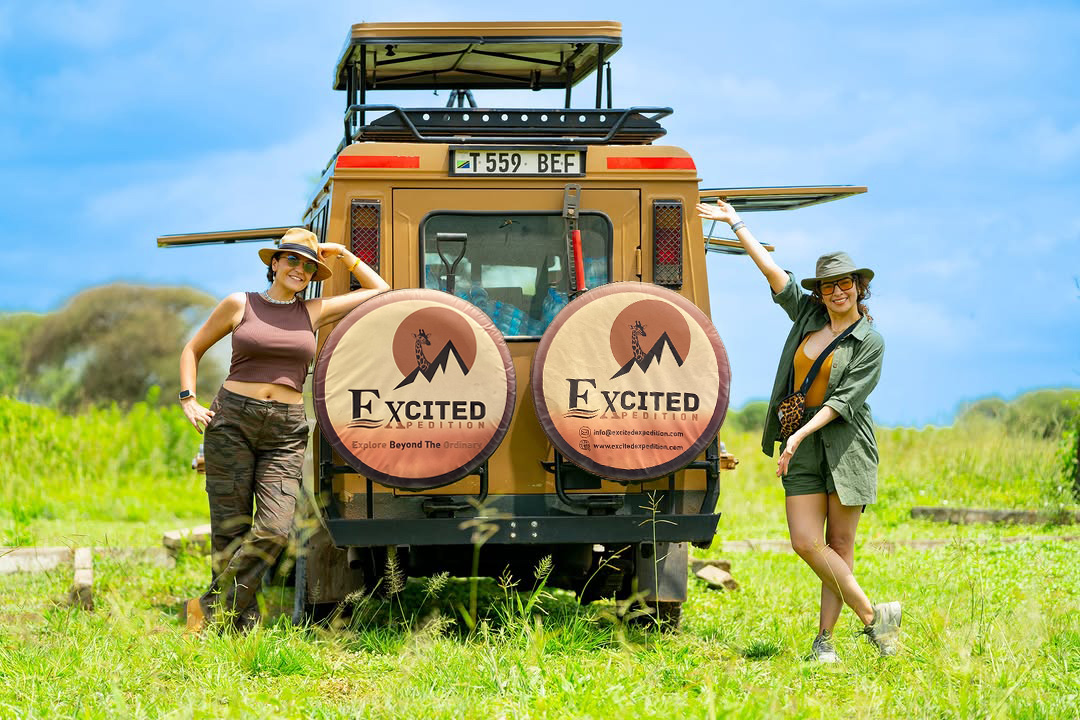 Two smiling women pose next to the back of a safari vehicle in a grassy field on a sunny day. The vehicle has two spare tires with the "Excited Expedition" logo.