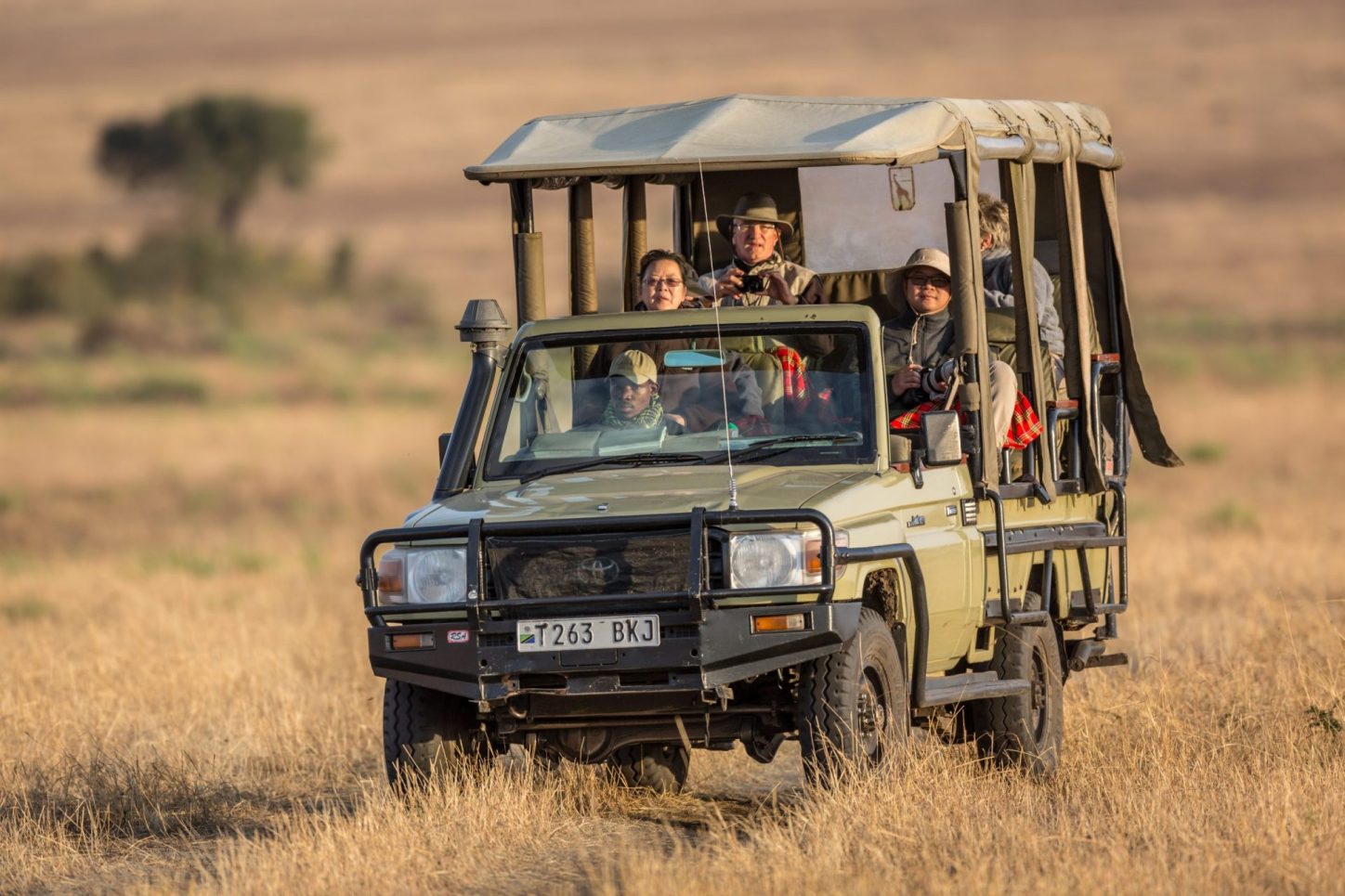A safari vehicle with tourists driving through a dry grassland, with passengers observing wildlife and taking photos.