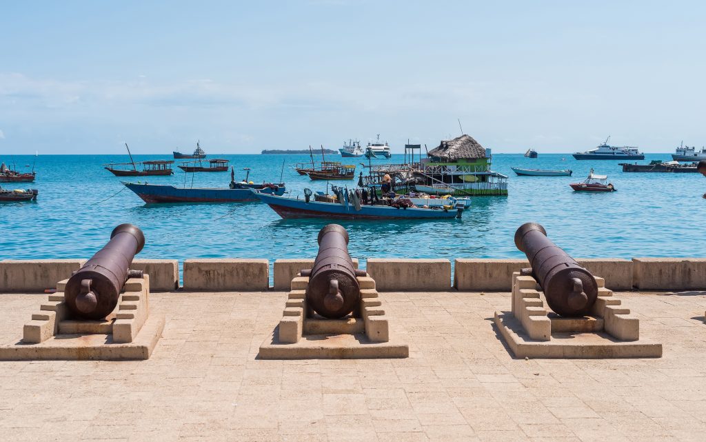 Old cannons facing the sea with traditional boats and floating structures on the blue waters of Zanzibar.