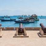 Old cannons facing the sea with traditional boats and floating structures on the blue waters of Zanzibar.