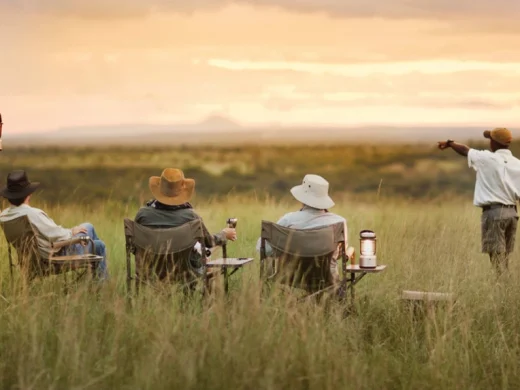 Three people in safari chairs and a guide enjoy a sunset view over an African plain.