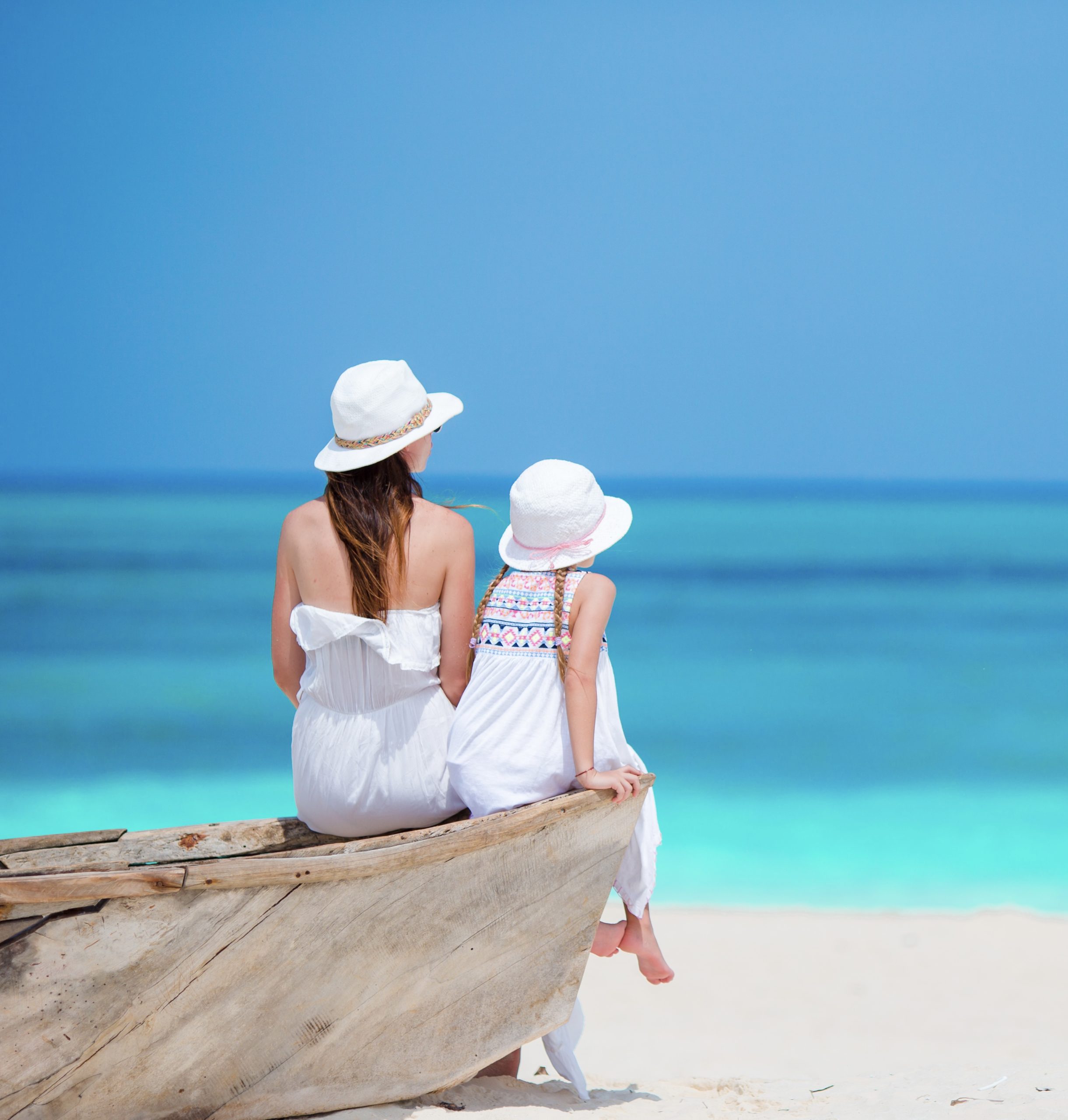 5 Days Discover Zanzibar Luxury Beach Holiday 1 A mother and daughter sitting on a rustic wooden boat on a stunning white sand beach, looking out at the bright turquoise ocean.