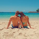 A couple sits in the sand at a tropical beach looking out at the turquoise ocean.